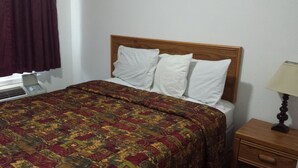 Ein Hotelzimmer mit einem hölzernen Kopfteil, einer gemusterten Tagesdecke, einem weißen Kissen, einem Nachttisch mit Lampe und einem Fenster mit Vorhang.
