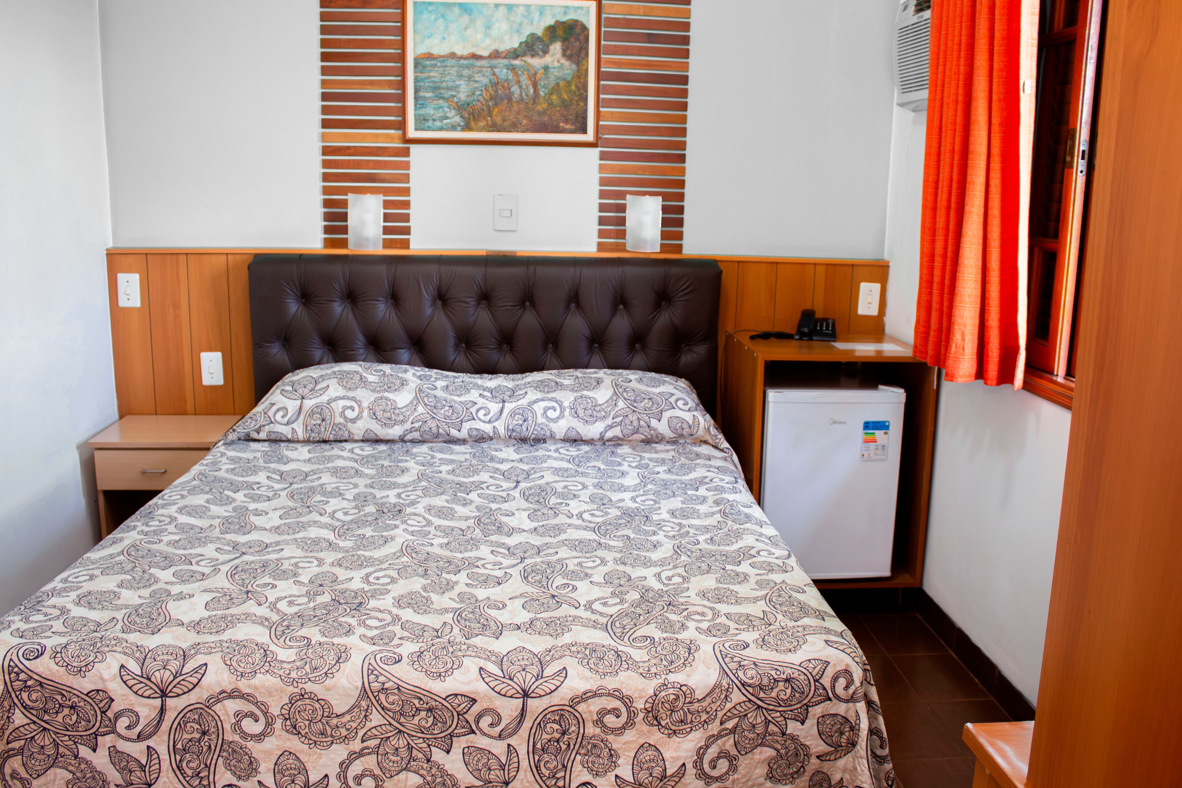 A bedroom with a bed, a nightstand, a small refrigerator, and a window with curtains.