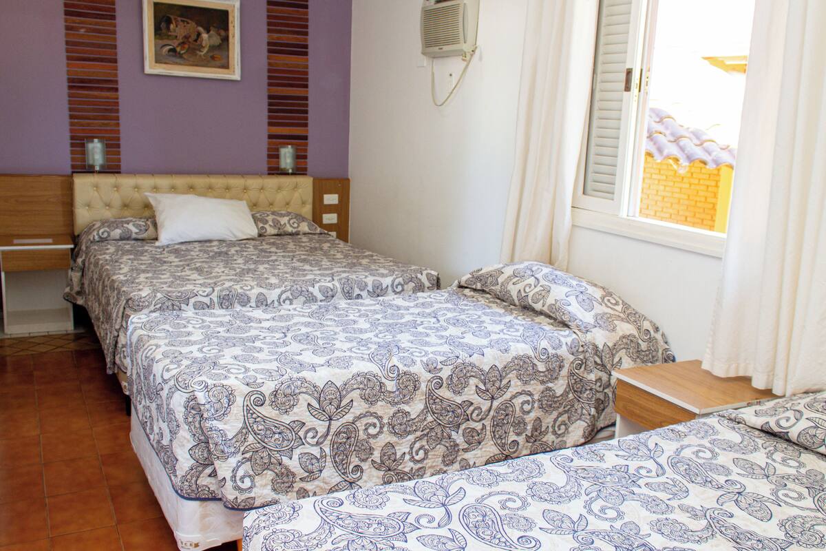 A bedroom with two beds, a wooden headboard, a bedside table, a window with curtains, and a wall-mounted air conditioner.