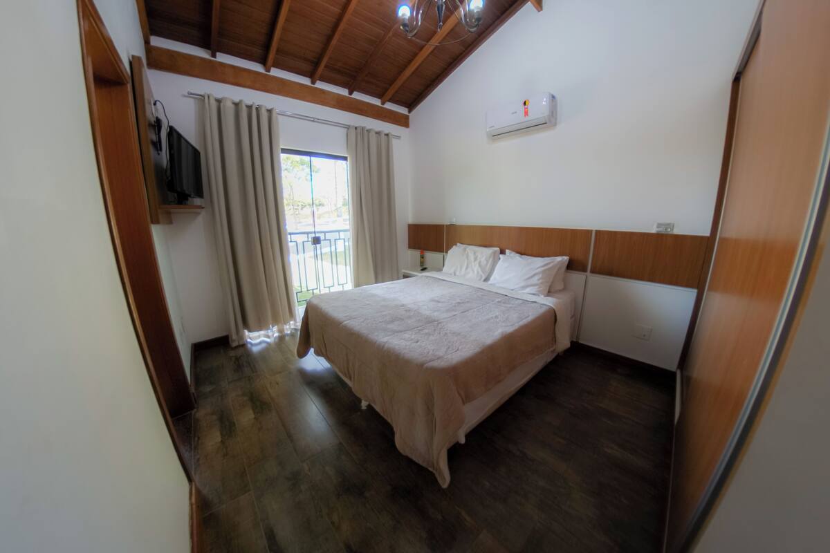 A bedroom with a wooden ceiling, a bed with a white headboard, a window with curtains, and an air conditioning unit on the wall.