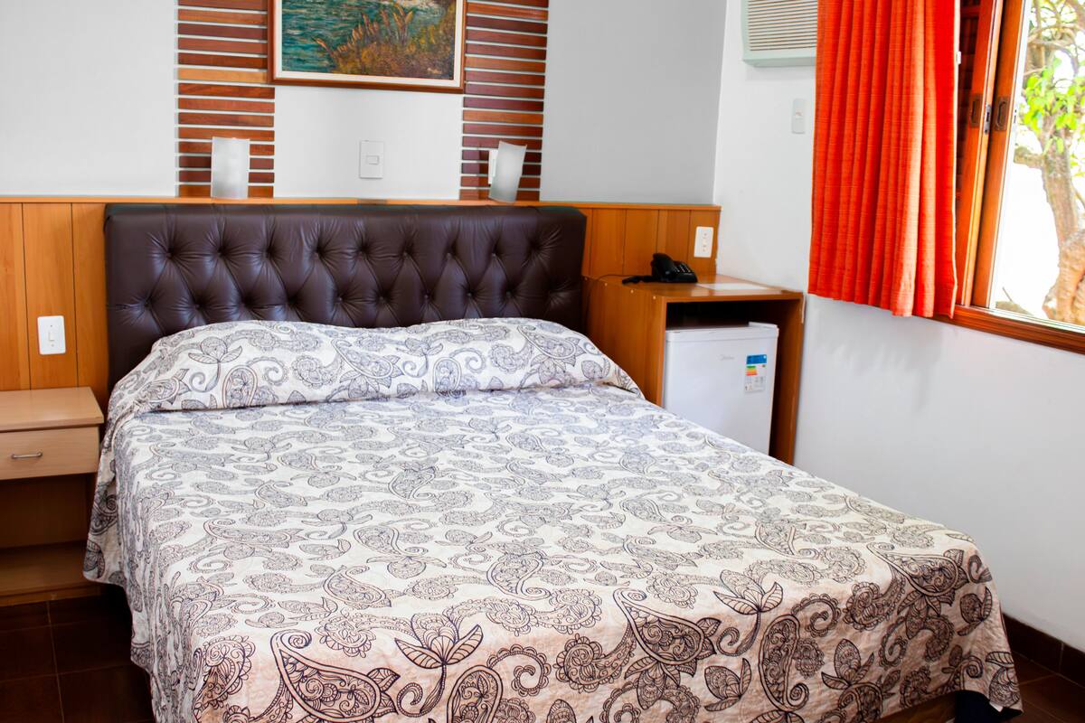 A neatly made bed with a patterned bedspread, a wooden headboard, a nightstand with a phone, and a small refrigerator.