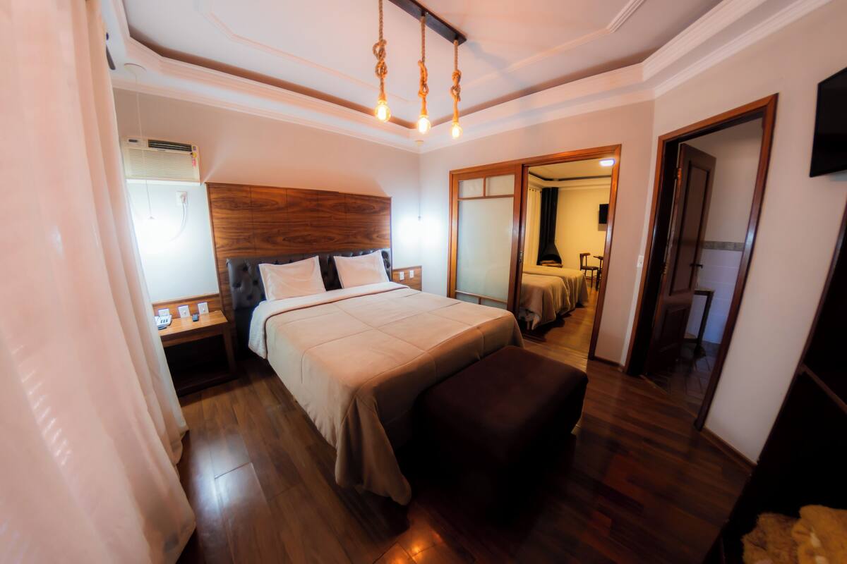 A bedroom with a wooden headboard, a large bed with a beige bedspread, a bedside table with a lamp, and a wooden floor.