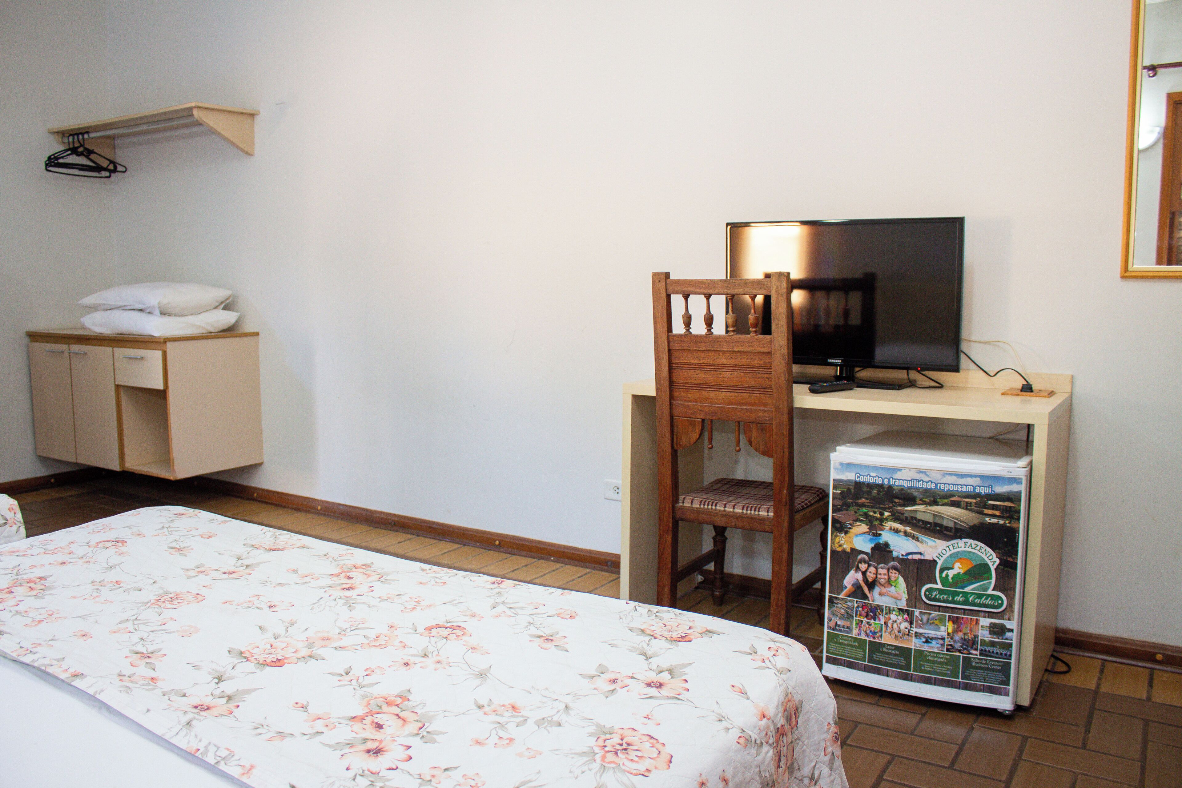 A hotel room with a bed, a desk with a computer, a chair, a TV, and a refrigerator.