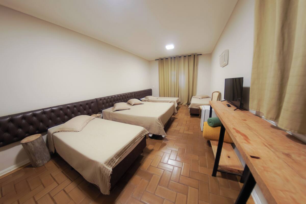 A hotel room with three beds, a wooden bench, a television, and a window with curtains.