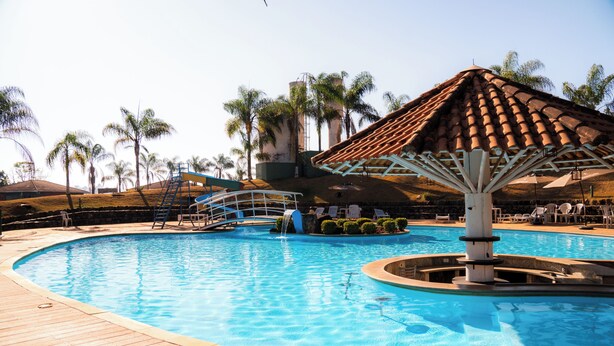 A swimming pool with a wooden deck, a water fountain, and a palm tree-lined background. - Hotel Fazenda Pocos de Caldas (Poços de Caldas)