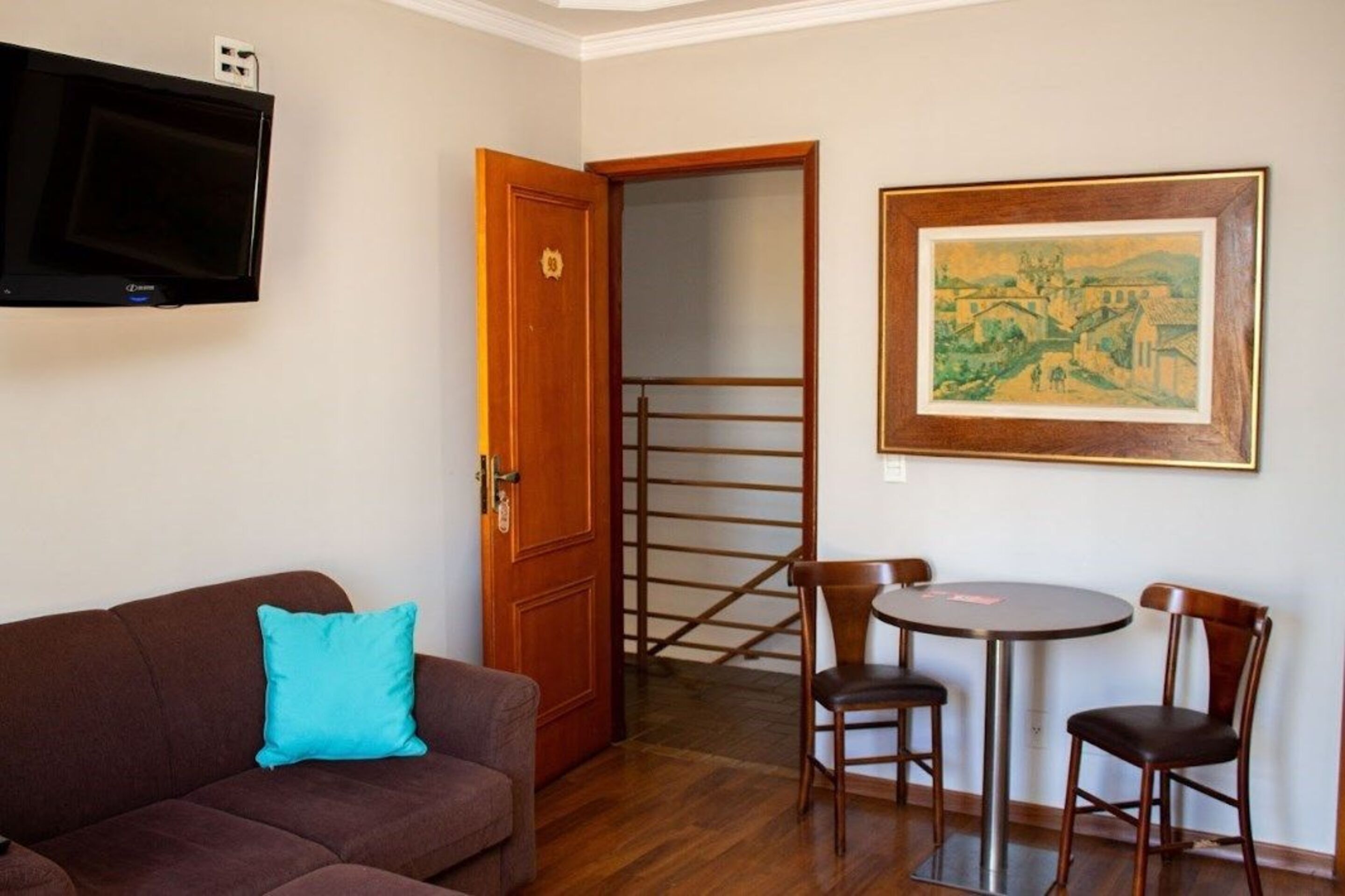 A room with a brown sofa, a teal pillow, a wooden door, a staircase, a framed painting, and a small round table with chairs.