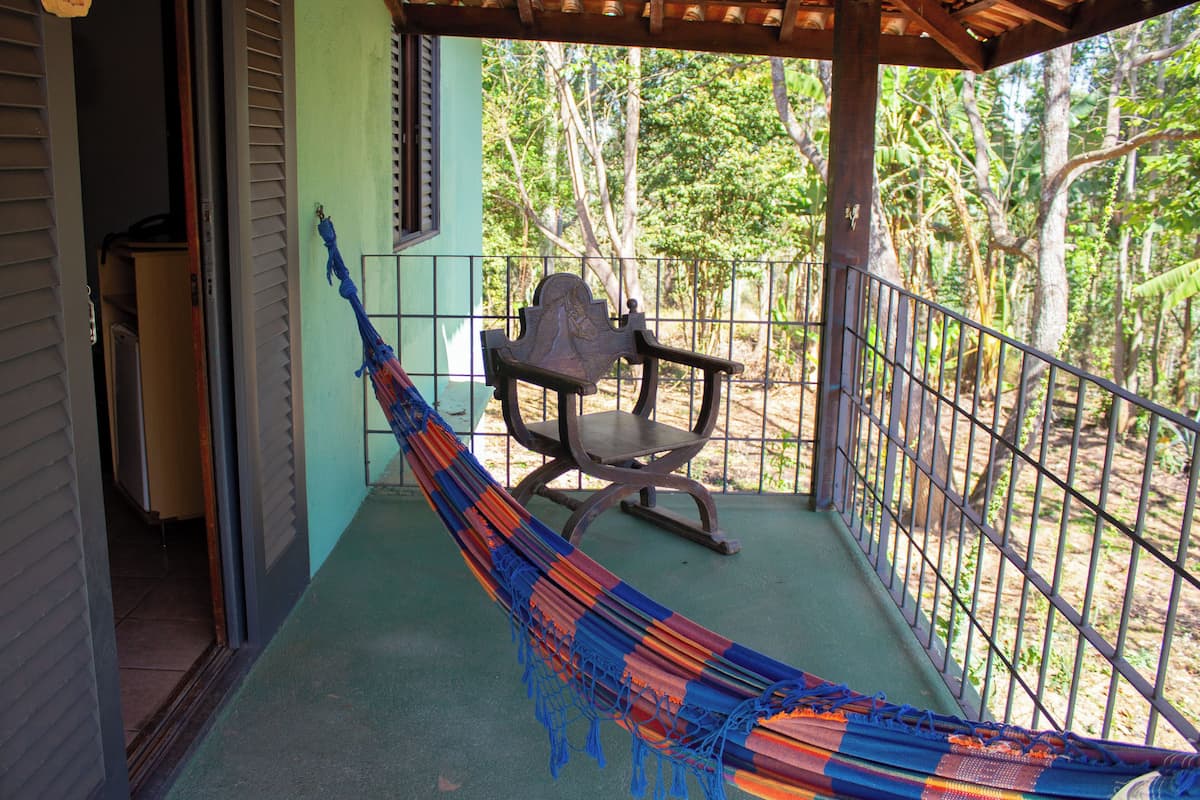 A hammock on a balcony with a chair, a view of trees, and a building in the background.