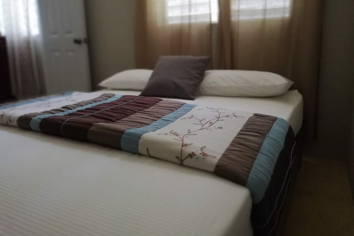 A neatly made bed with a quilt and pillows, a window with curtains, and a door in the background.