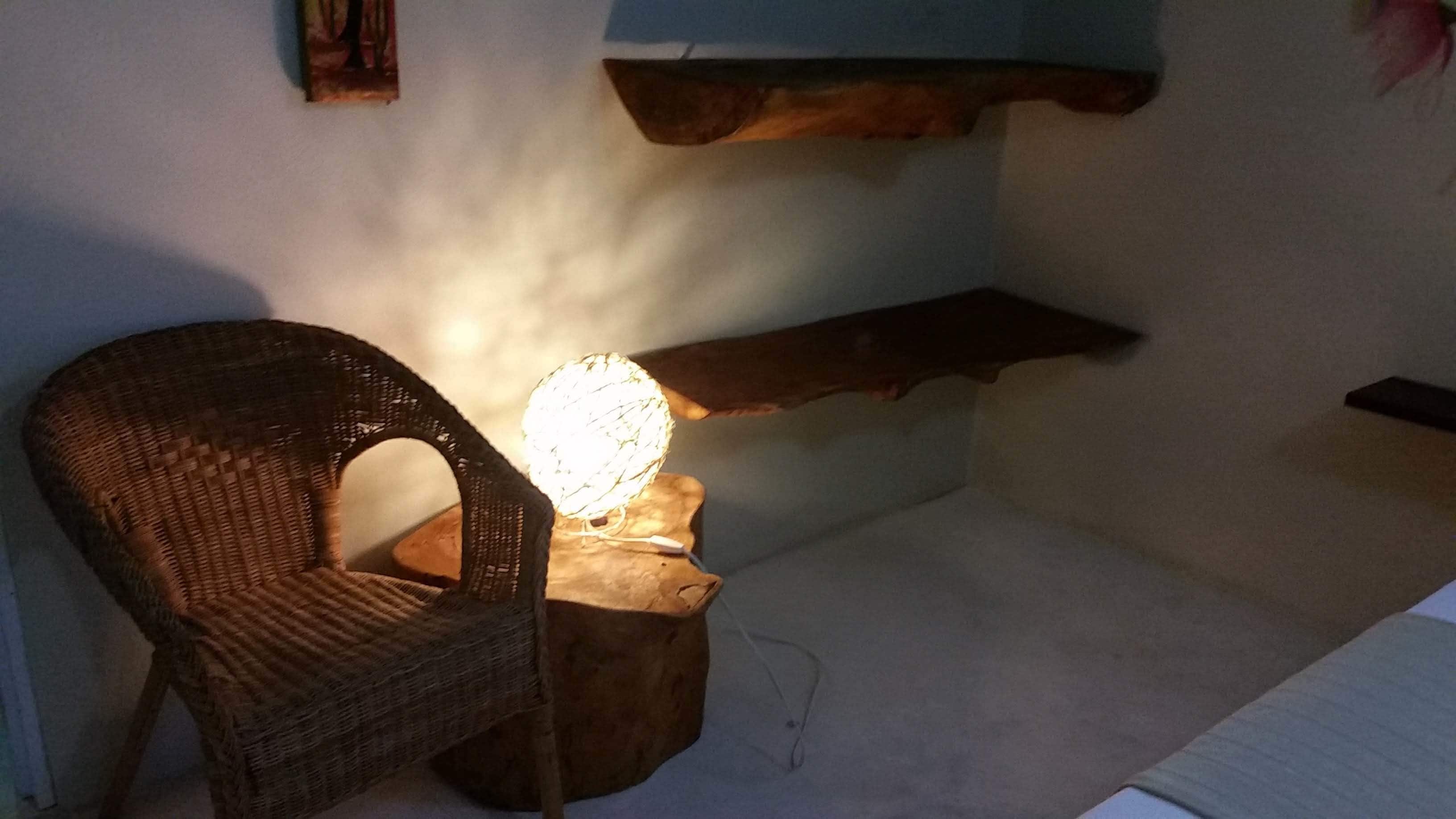 A wicker chair with a light on a wooden stool, a shelf with a picture, and a wall with a textured finish.