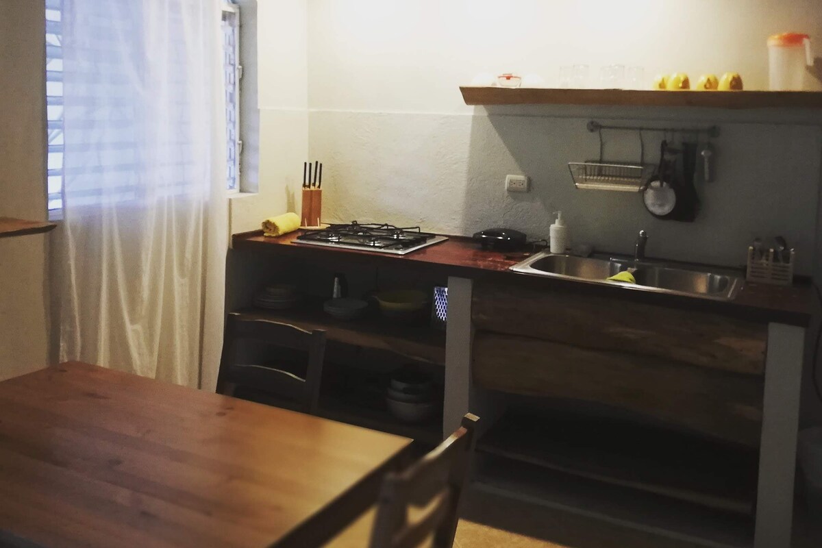 Une cuisine compacte avec une table et des chaises en bois, une cuisinière, un évier et une fenêtre agrémentée d’un rideau blanc.