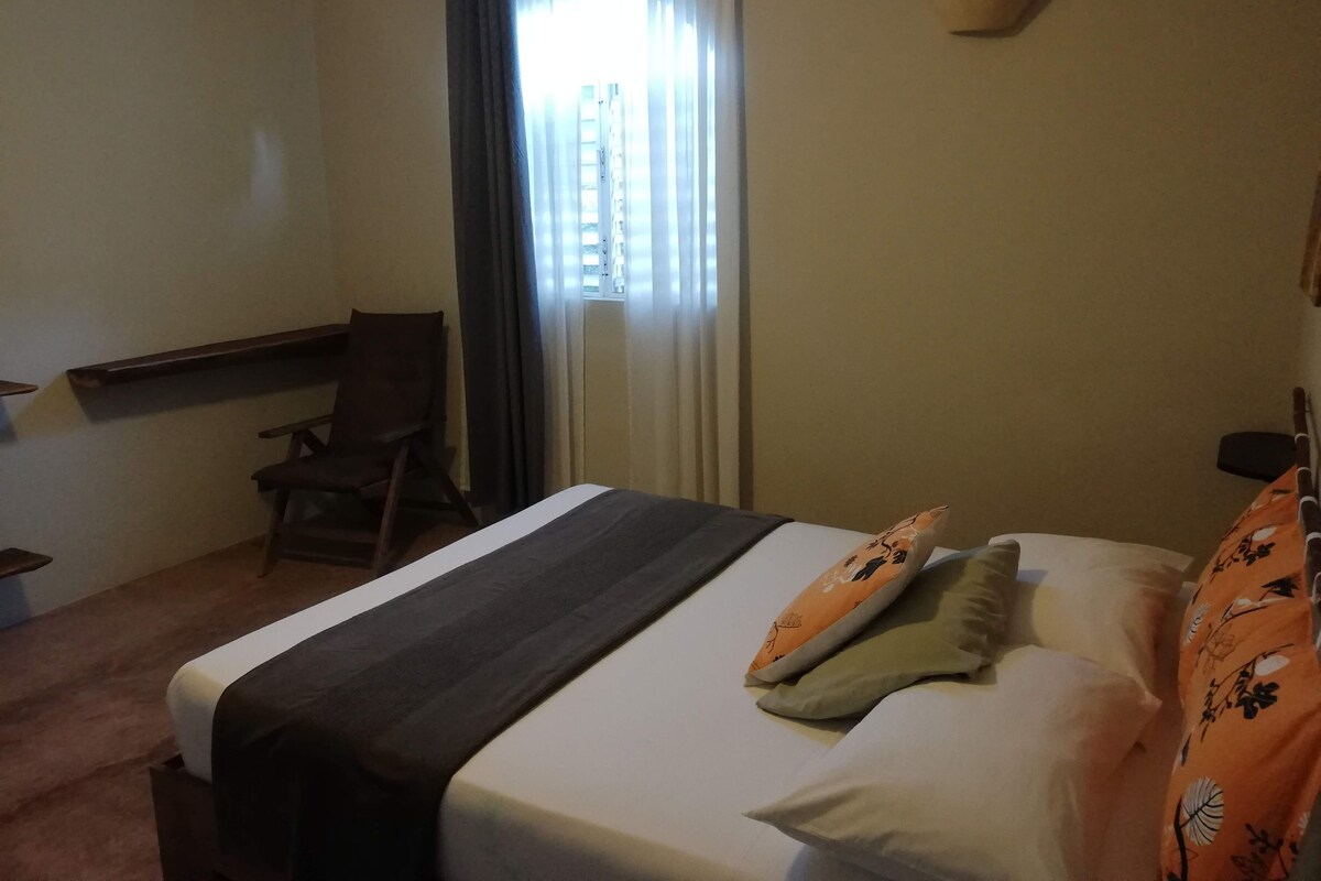 Una habitación de hotel con cama, una silla, un ventanal con cortinas y un equipo de aire acondicionado.