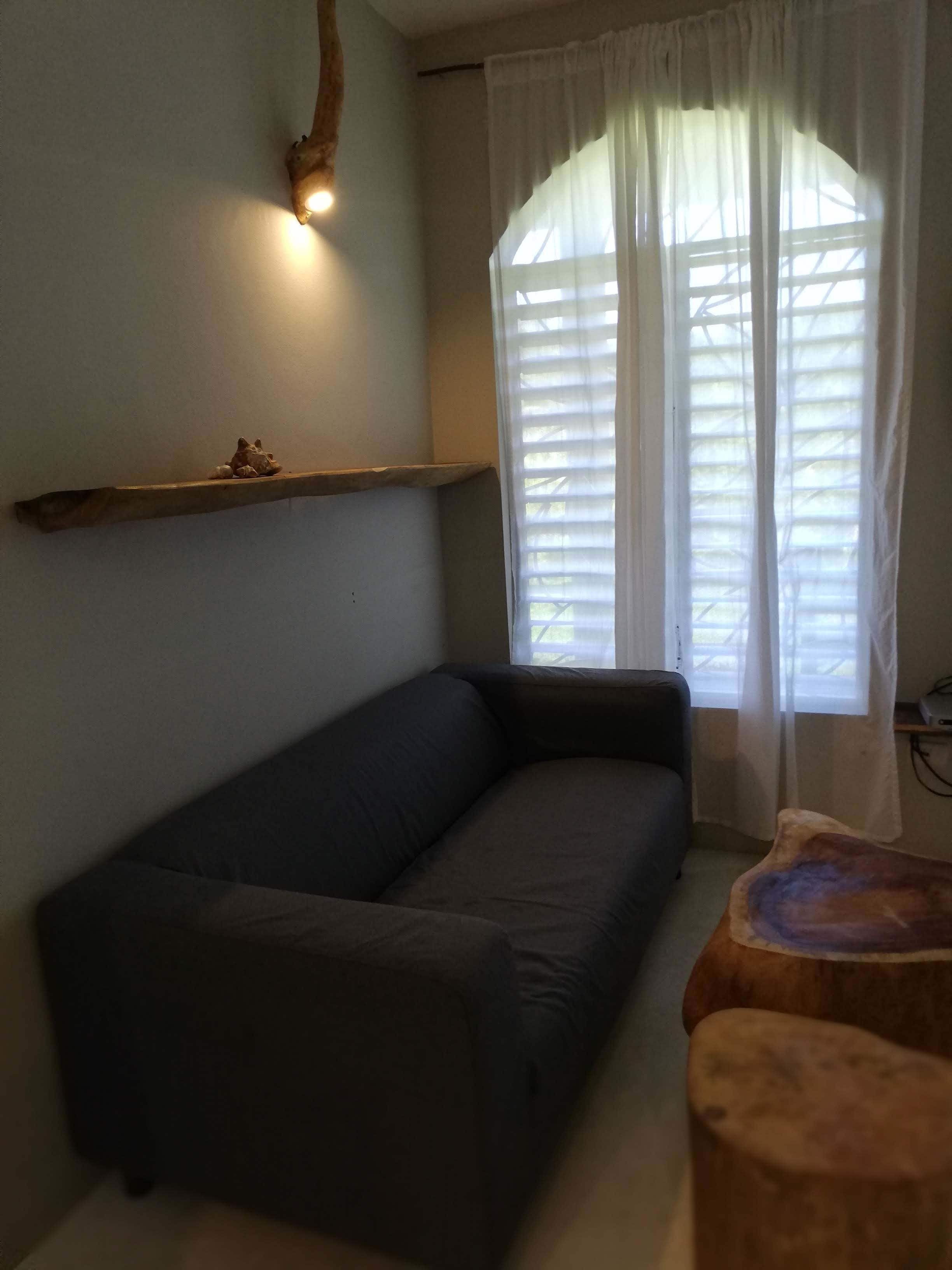 A living room with a dark sofa, a wooden shelf, a window with sheer curtains, and a small wooden stool.