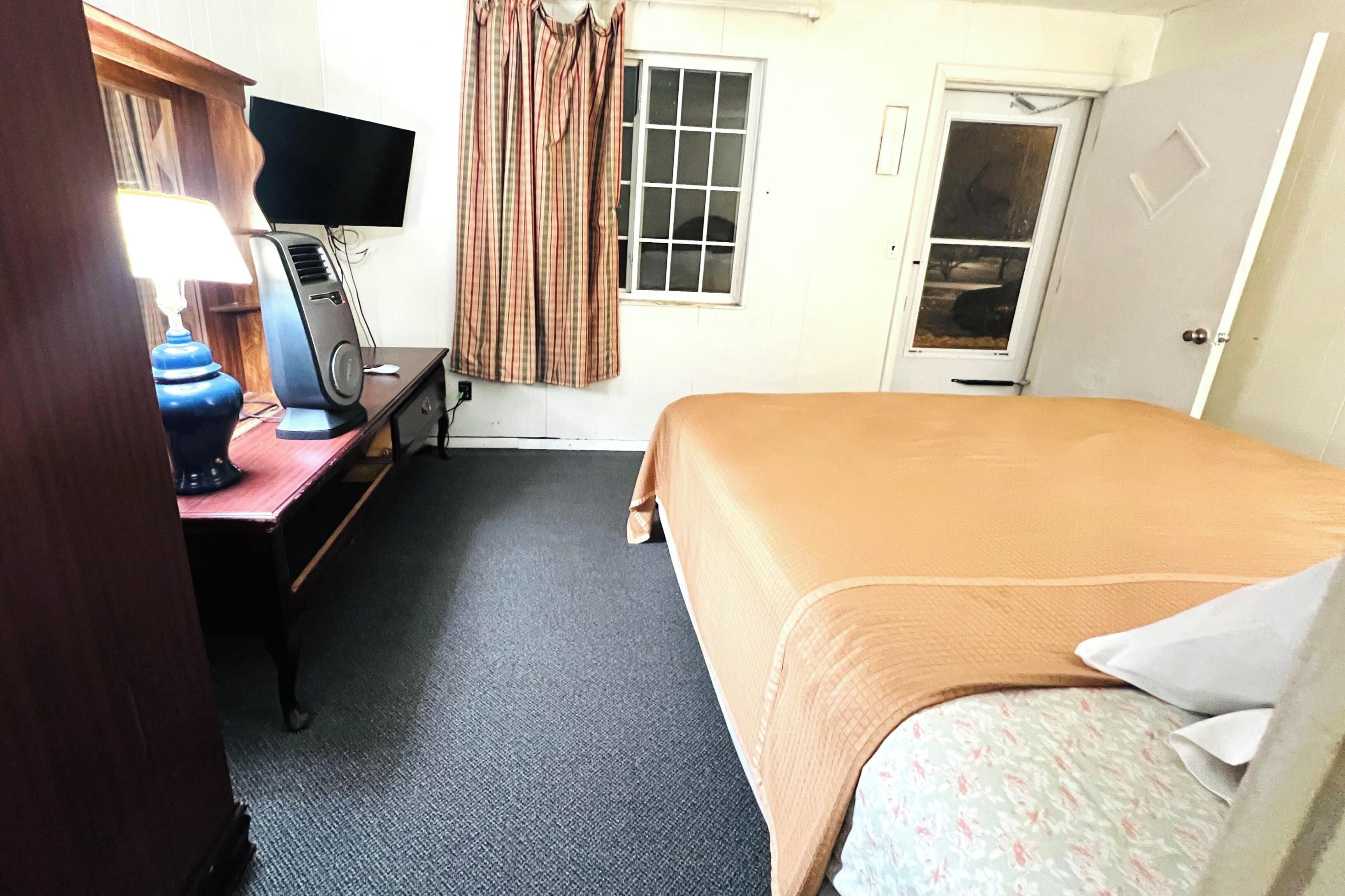 Una camera d'albergo con un letto, una scrivania con computer e lampada, una finestra con tende e una porta.