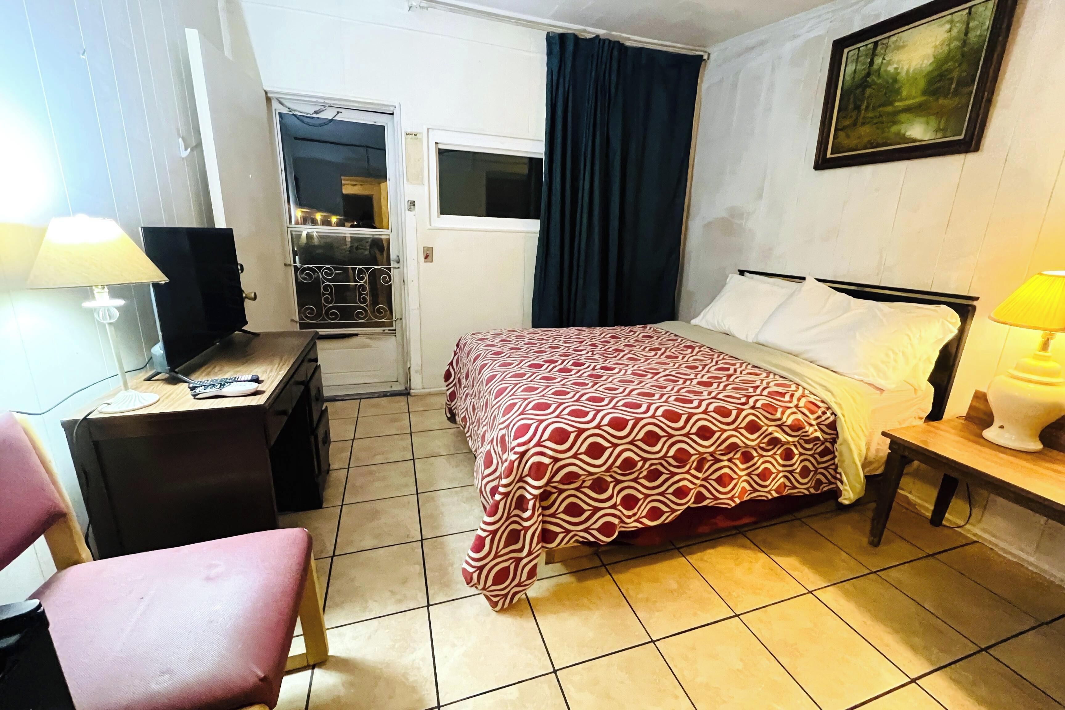 A hotel room with a bed, a desk with a lamp, a chair, a wardrobe, and a painting on the wall.