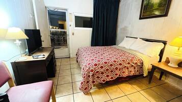 A hotel room with a bed, a desk with a lamp, a chair, a wardrobe, and a painting on the wall.