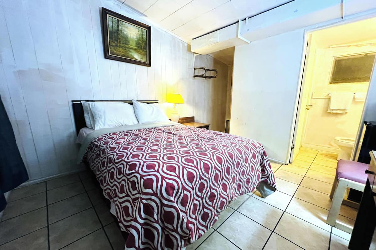A bedroom with a bed, a nightstand, a lamp, a door leading to a bathroom, and a painting on the wall.