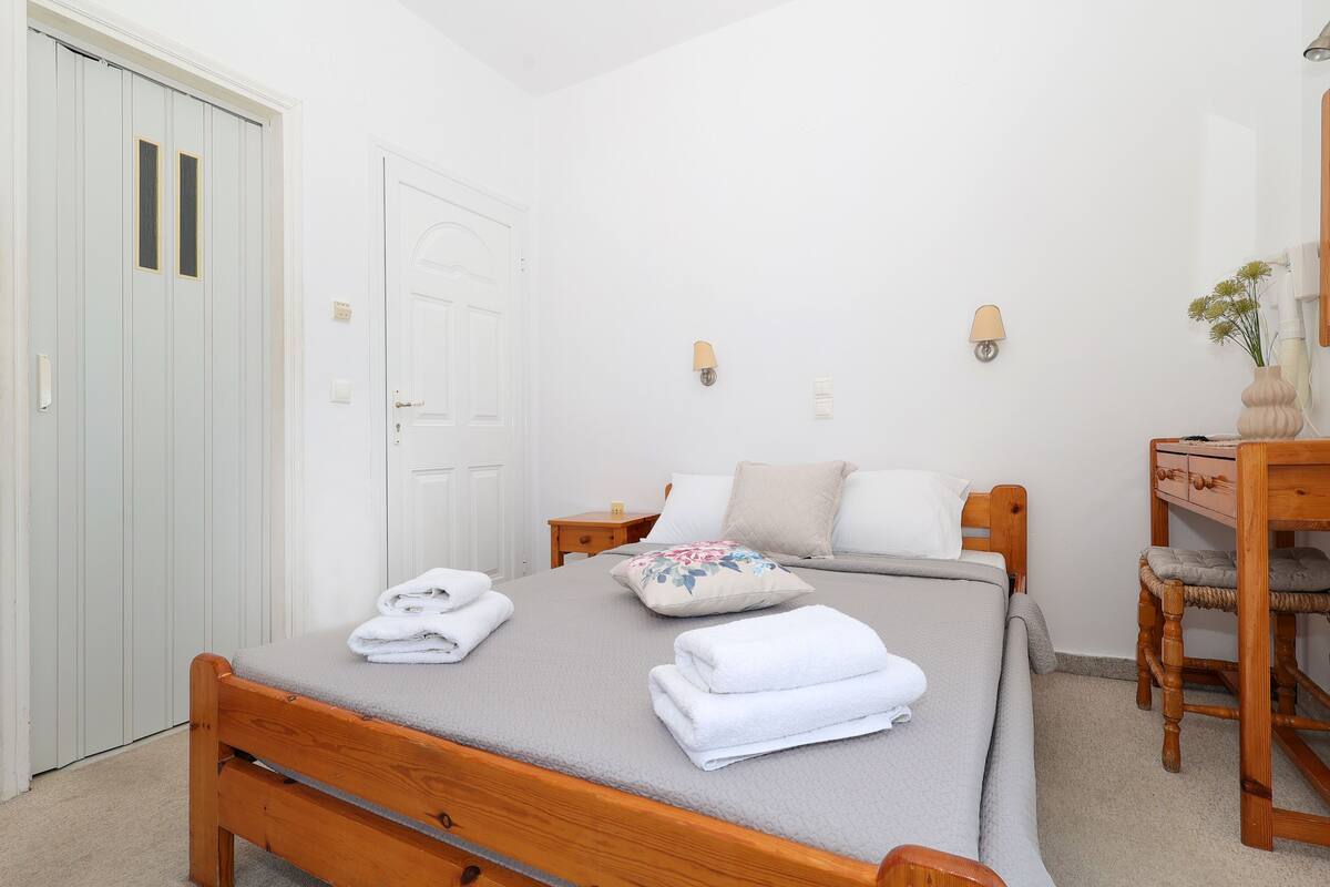 A neatly made bed with folded towels, a wooden nightstand, a chair, and a desk with a vase of flowers.