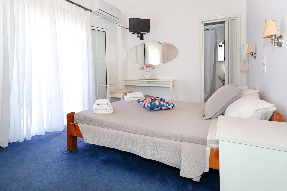 A bedroom with a bed, a bedside table, a mirror, a TV, and a bathroom door.