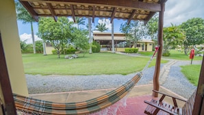 Una hamaca bajo una pérgola de madera, un banco de madera y una vista de un césped cuidado con árboles y un edificio al fondo.