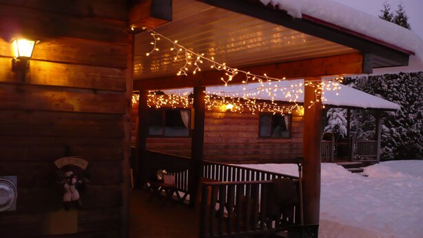 A wooden cabin with a porch decorated with string lights, a covered seating area, and a snow-covered roof. - Shames Country Lodge (Terrace)