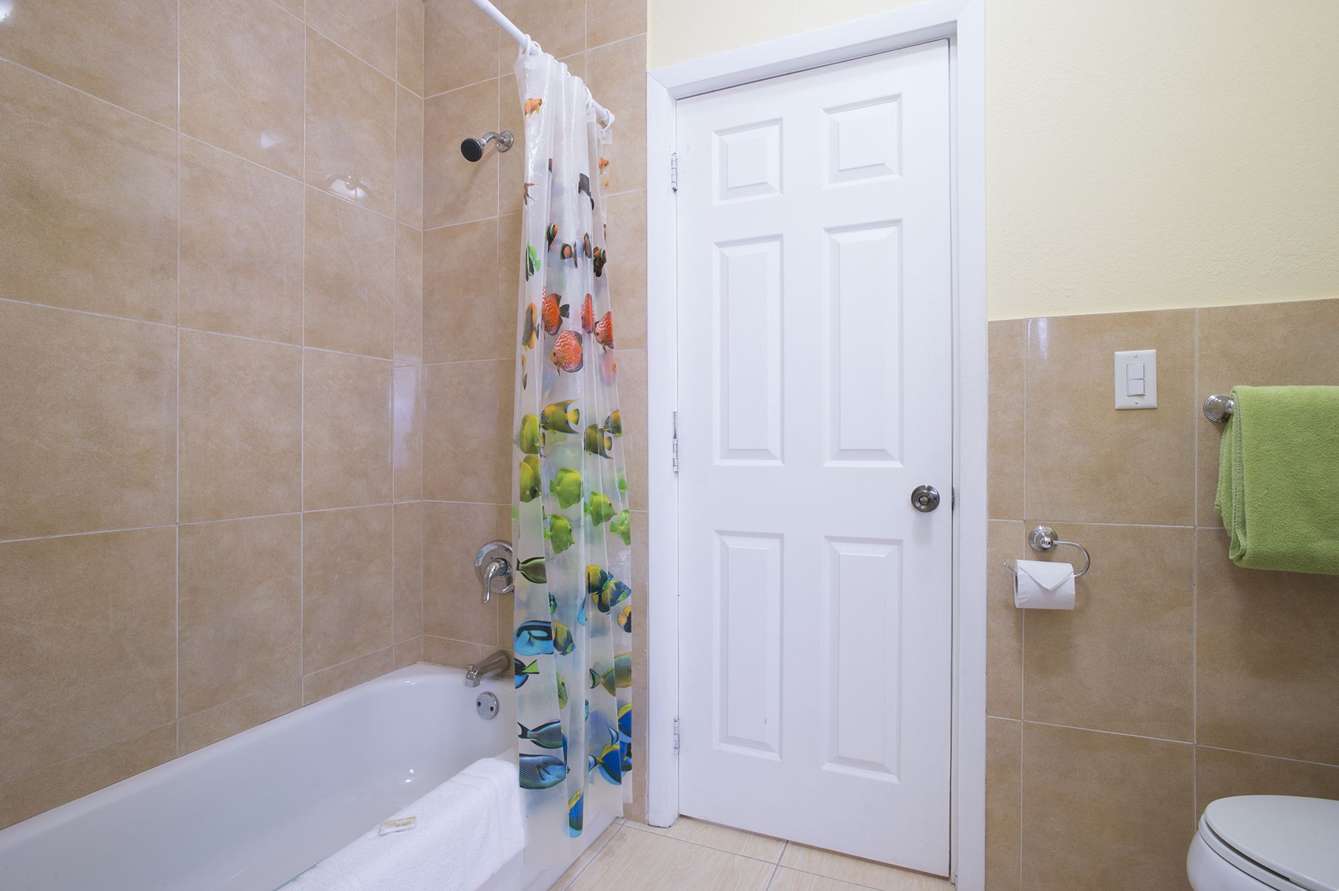 Un baño con bañera, cortina de ducha con motivos de peces, una puerta blanca y una toalla verde en la pared.