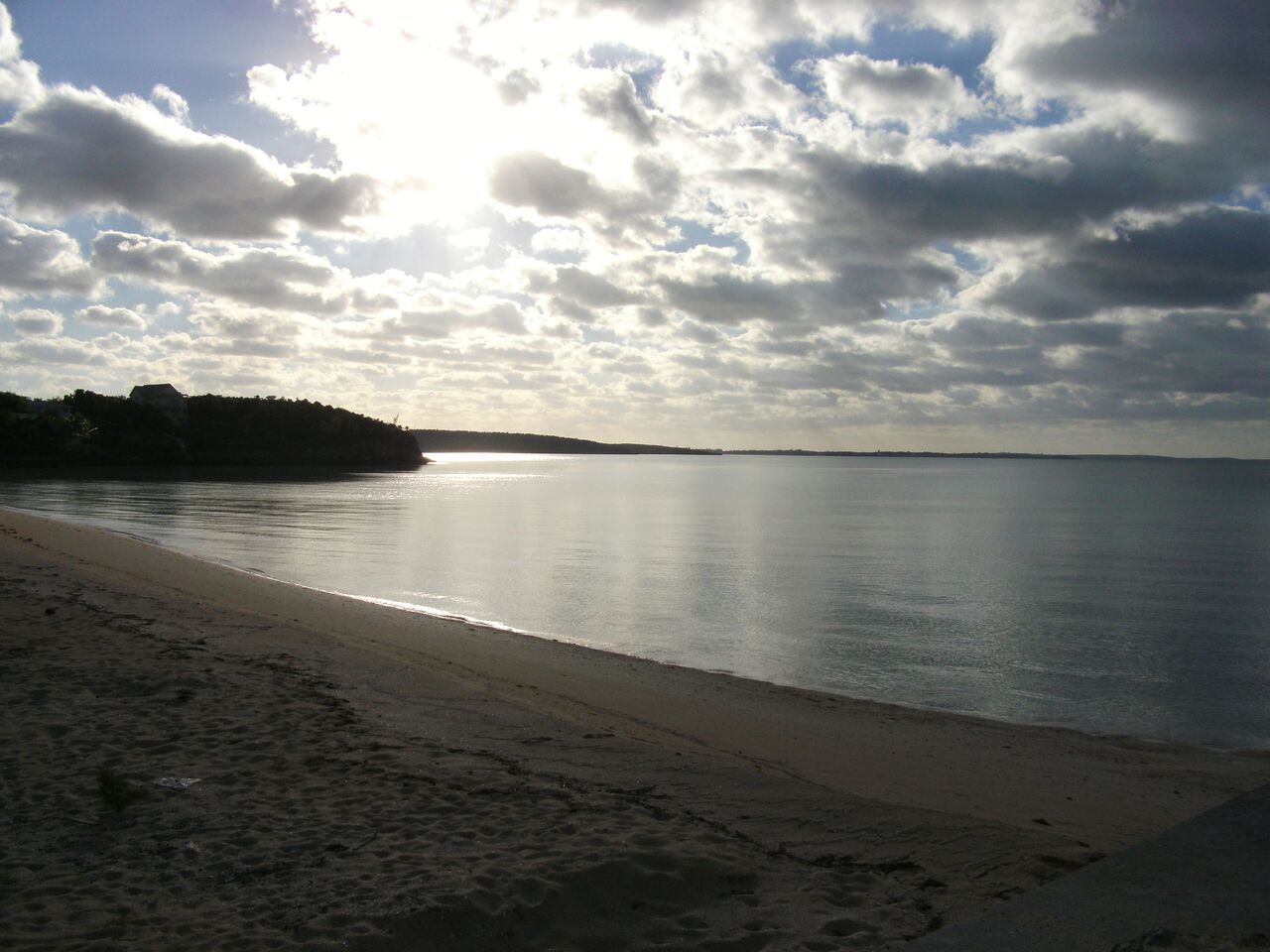 Ein Strand mit ruhiger See, sandigem Ufer und einer entfernten Landmasse unter teilweise bewölktem Himmel.