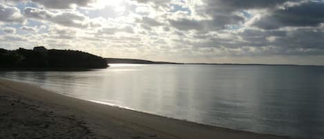 Ein Strand mit ruhiger See, sandigem Ufer und einer entfernten Landmasse unter teilweise bewölktem Himmel.