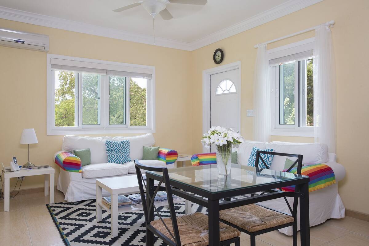 A bright living room with a glass-top dining table, white sofas with colorful pillows, a small white side table, and a black woven chair.