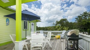 Una terraza de madera con sillas blancas, una mesa, una parrilla y un paraguas azul.