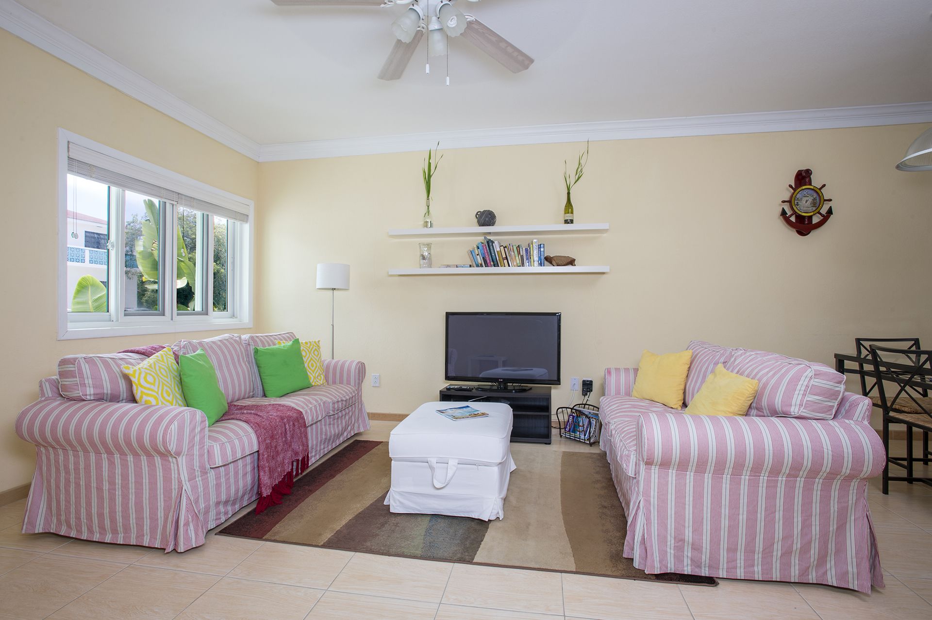 A living room with two striped sofas, a white ottoman, a flat-screen TV, and a ceiling fan.