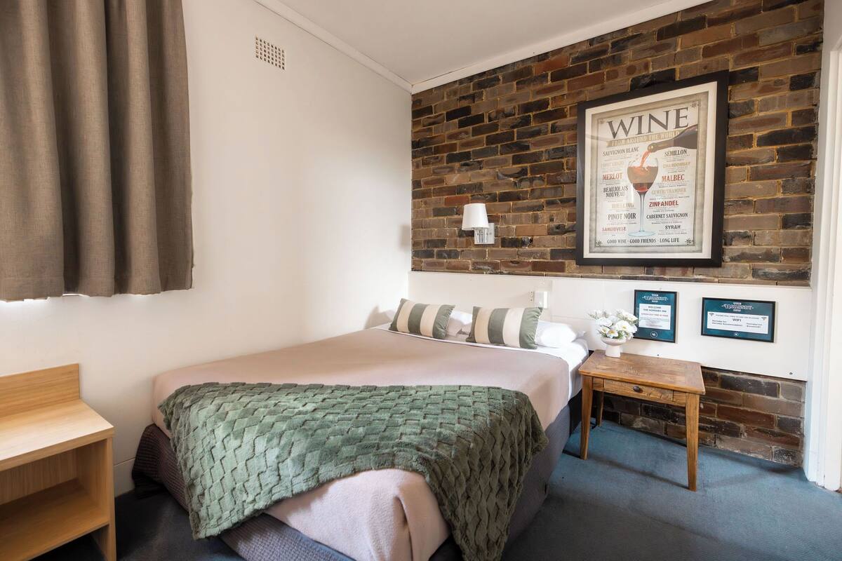 A hotel room with a brick wall, a bed with a green blanket, a wooden nightstand, and a framed wine poster.