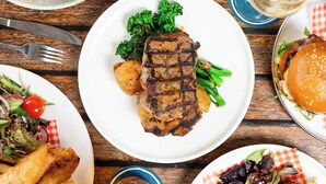 Ein Esstisch mit einer Auswahl an Speisen, darunter gegrilltes Steak, Kartoffeln, Brokkoli und ein Burger.