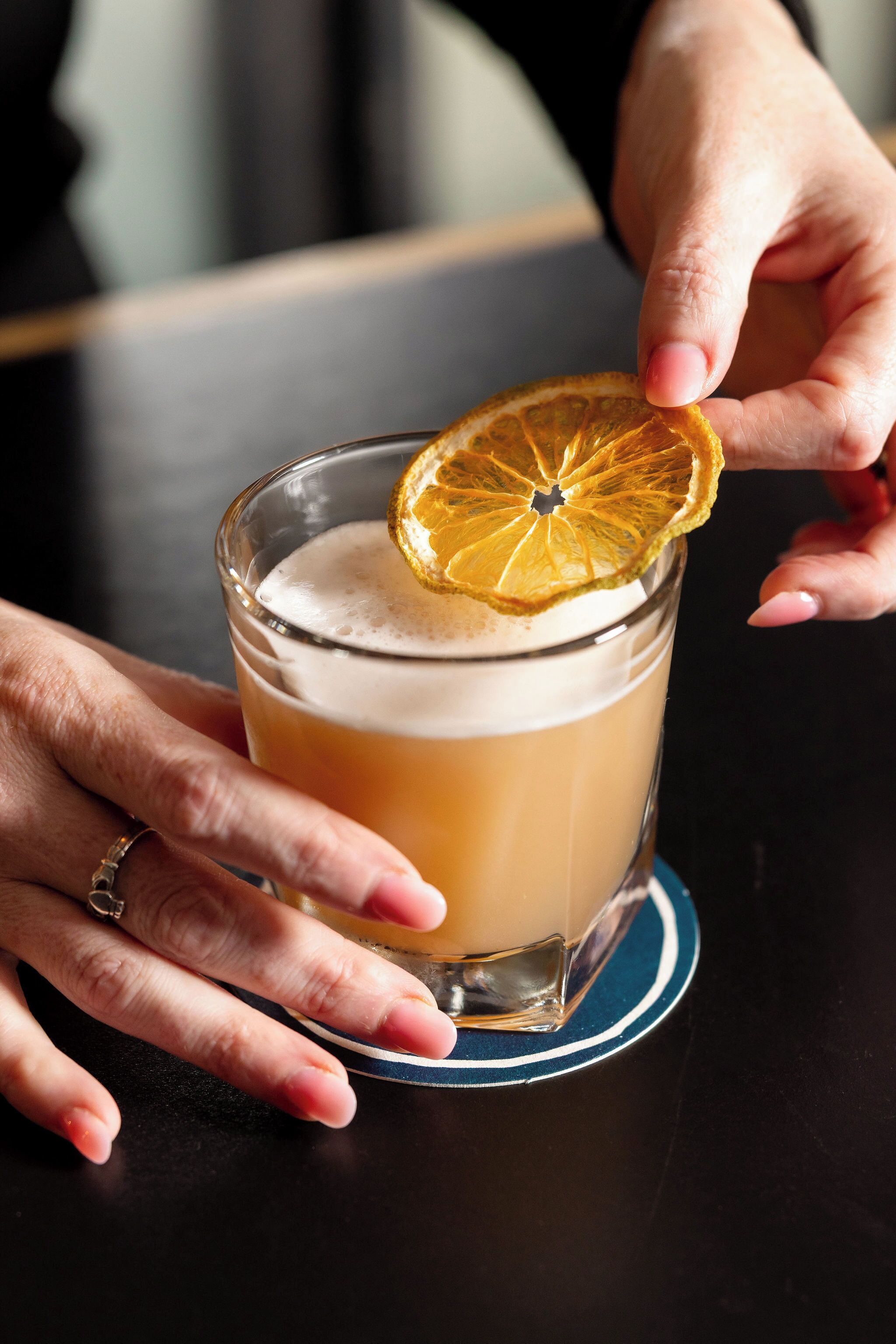 A hand holding a glass with a slice of orange garnish on a coaster.