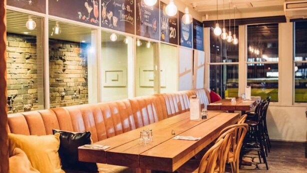 Ein Restaurant mit einem langen Holztisch, gepolsterten Sitzplätzen, Korbstühlen und einer Ziegelwand.
