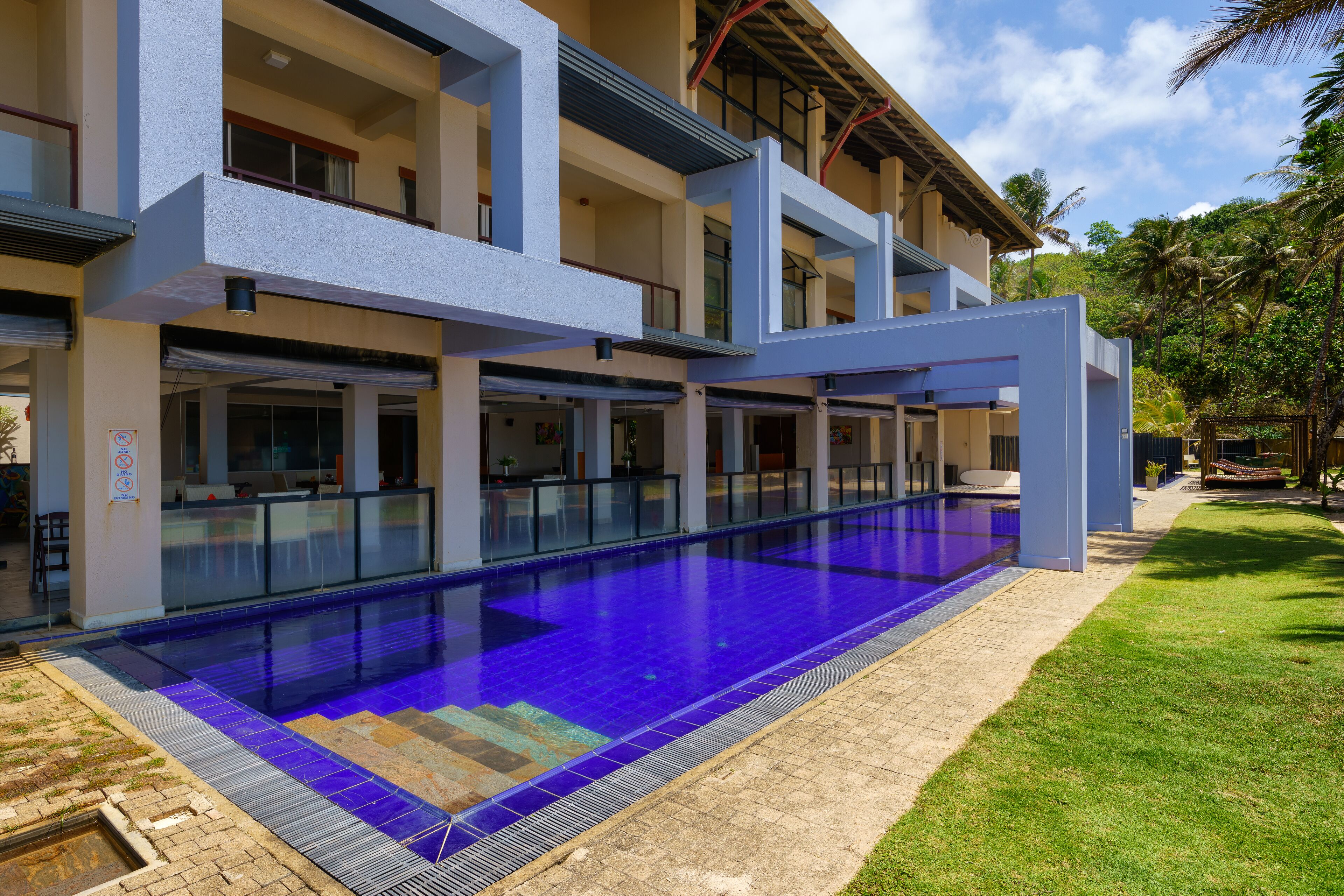 A modern hotel with a large outdoor swimming pool, multiple balconies, and a well-maintained lawn.