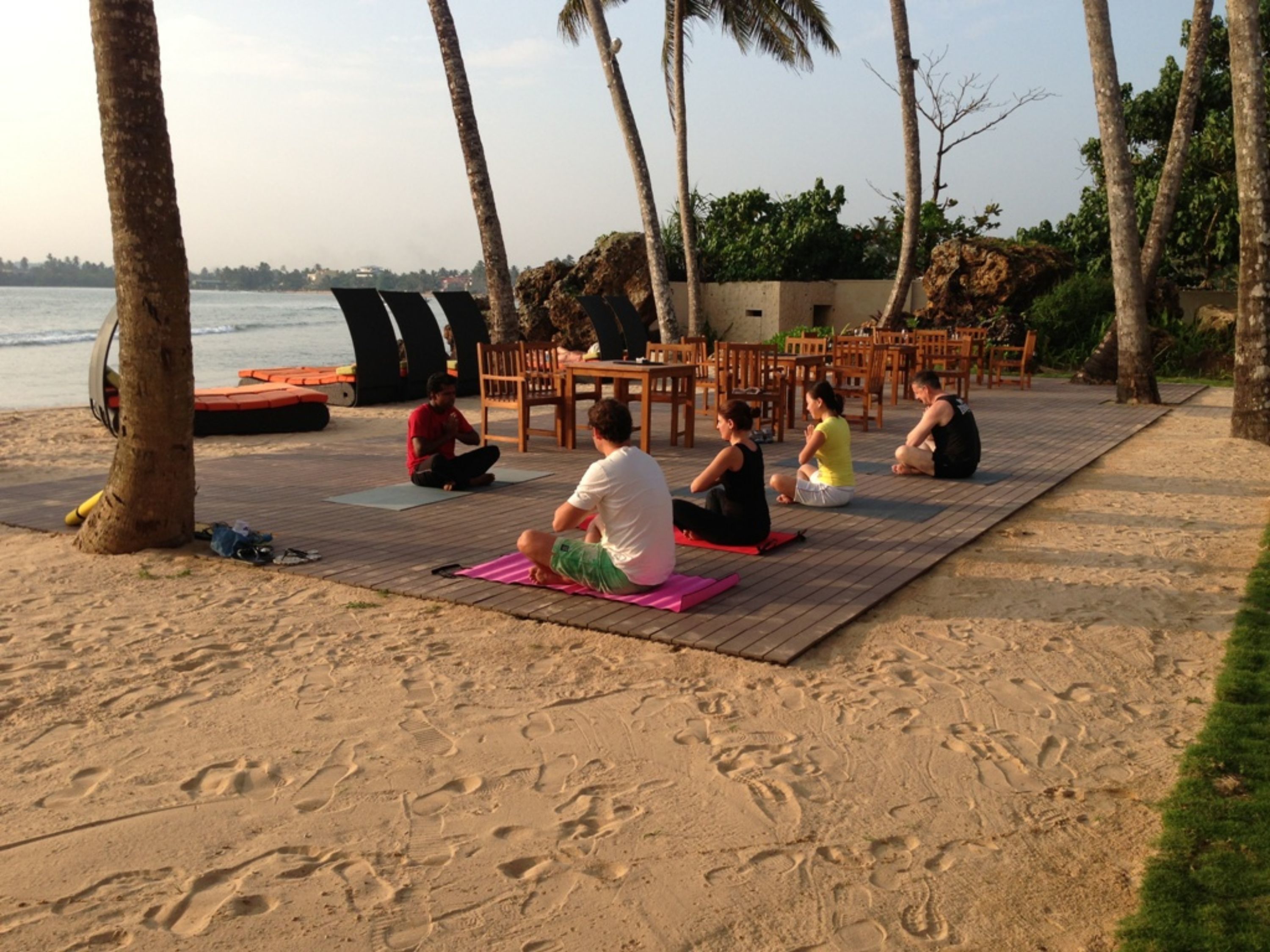 Clase de yoga en la playa con participantes sentados en esterillas, rodeados de mobiliario de madera y palmeras.