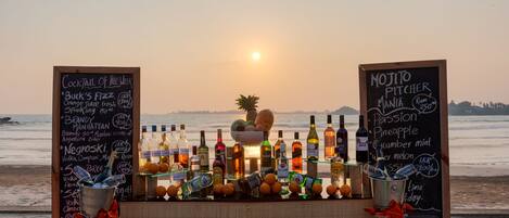 A beachside bar with a sunset view, featuring cocktail and mojito menus, and a variety of bottles and fruits.