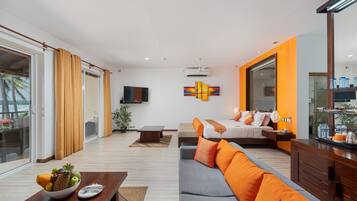 A modern living room with a sofa, dining table, and a fruit bowl. There is a bed with orange bedding and a TV mounted on the wall.
