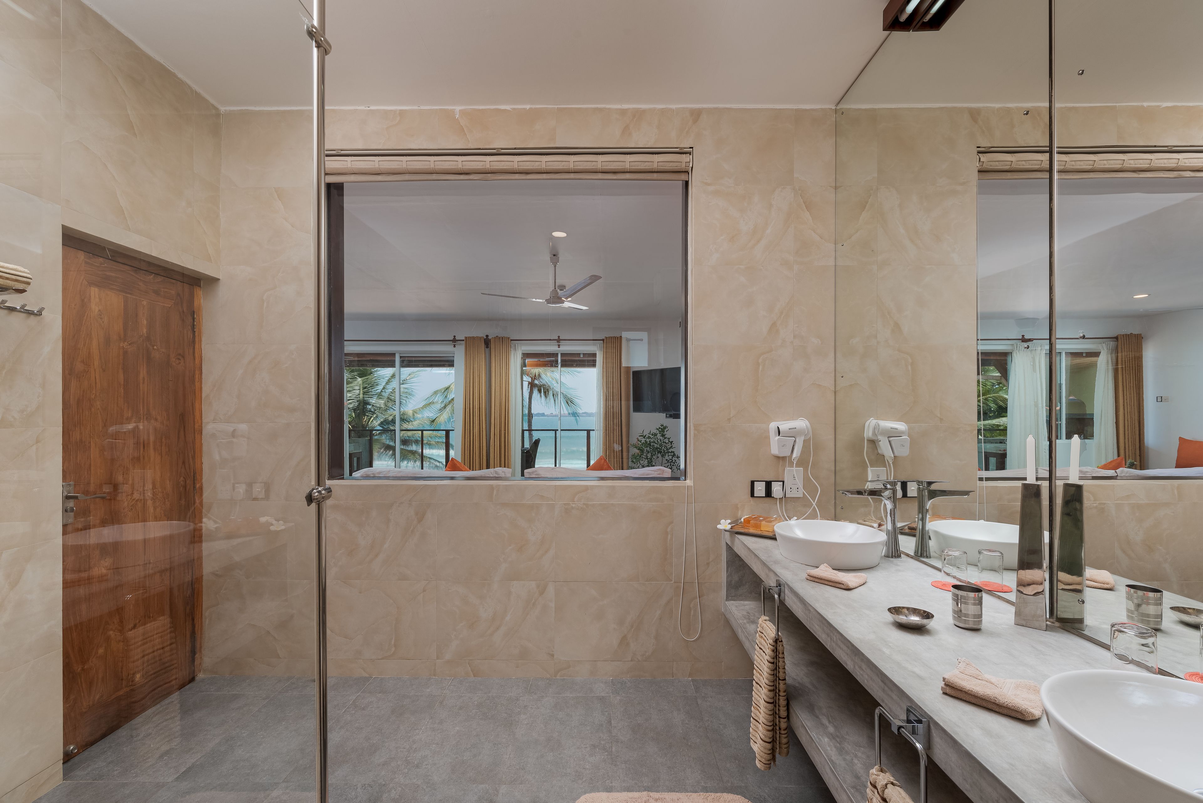 A modern bathroom with a large mirror, two sinks, a shower, and a wooden door.