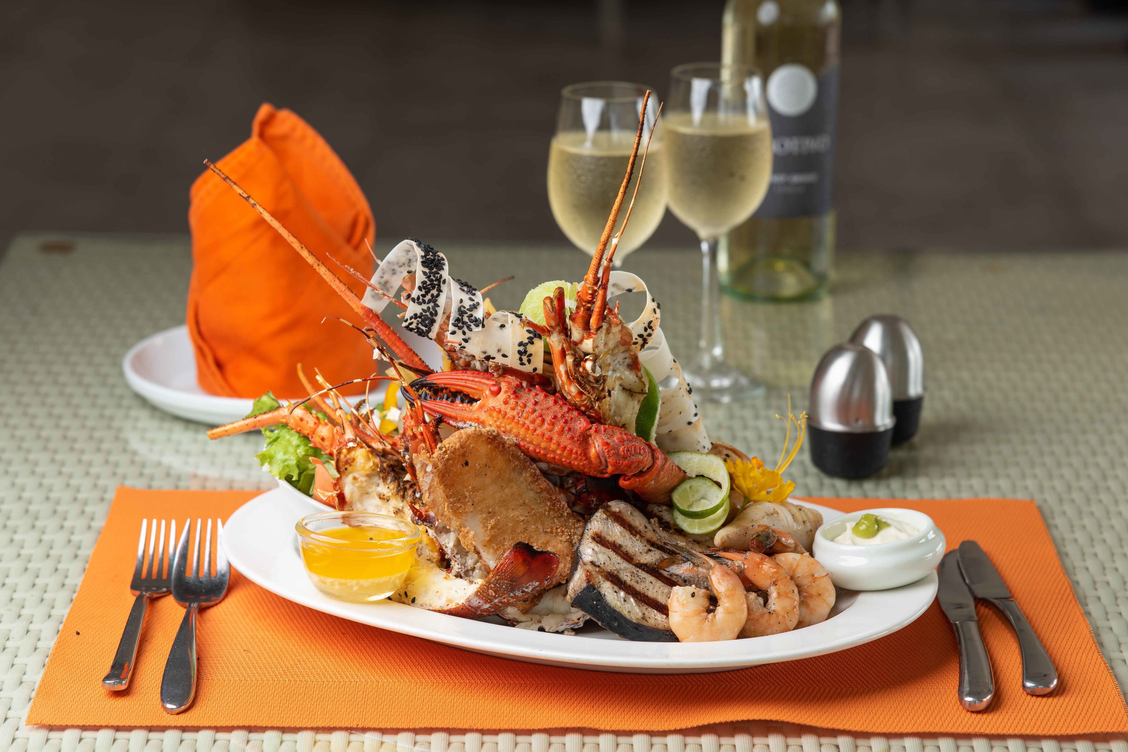 Un plateau de fruits de mer comprenant divers mets grillés, un verre de vin blanc et une bouteille de vin, le tout posé sur une table recouverte d’un set de table en fibres tressées.