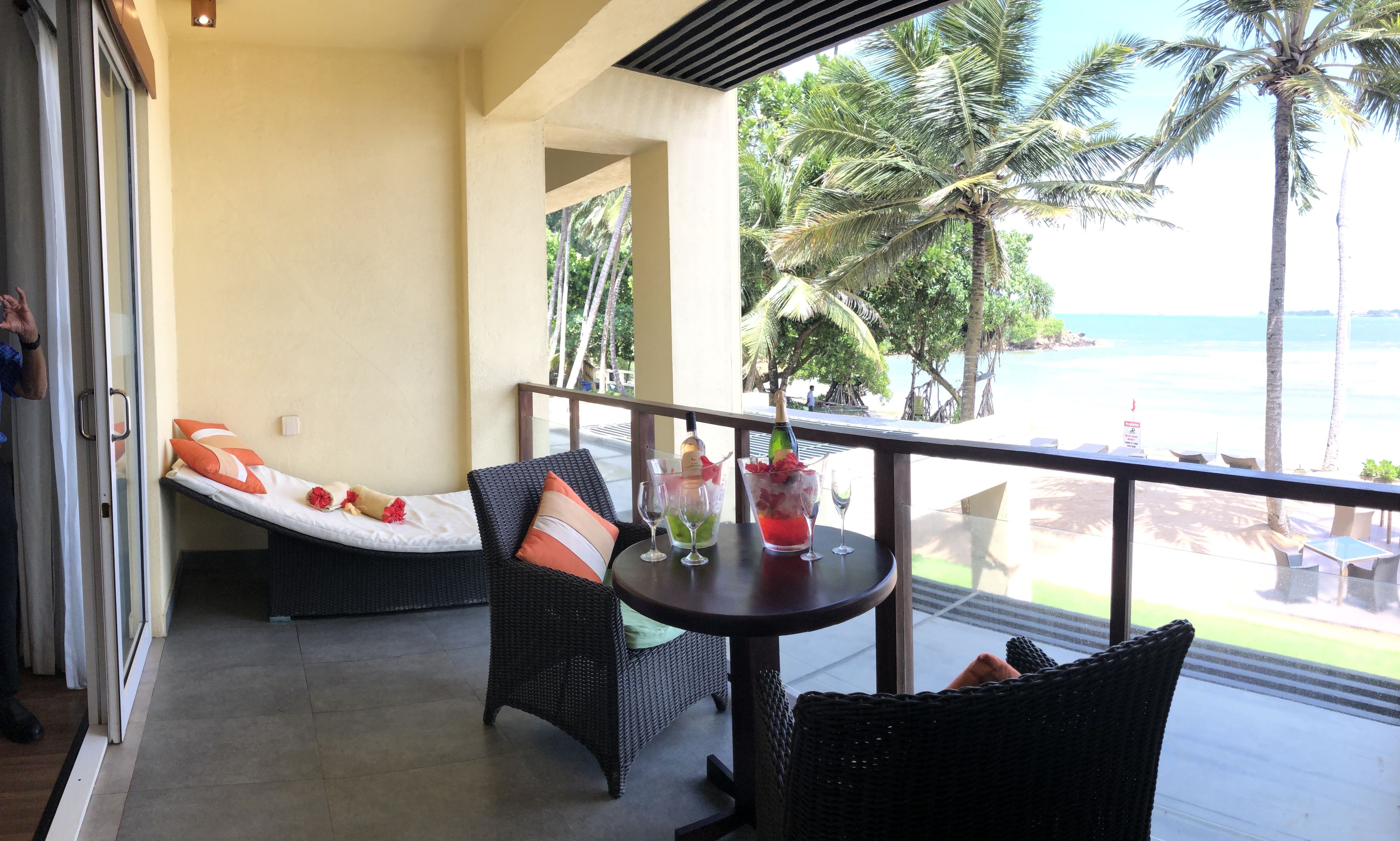 A balcony with wicker furniture, a table with drinks, and a view of the beach and palm trees.