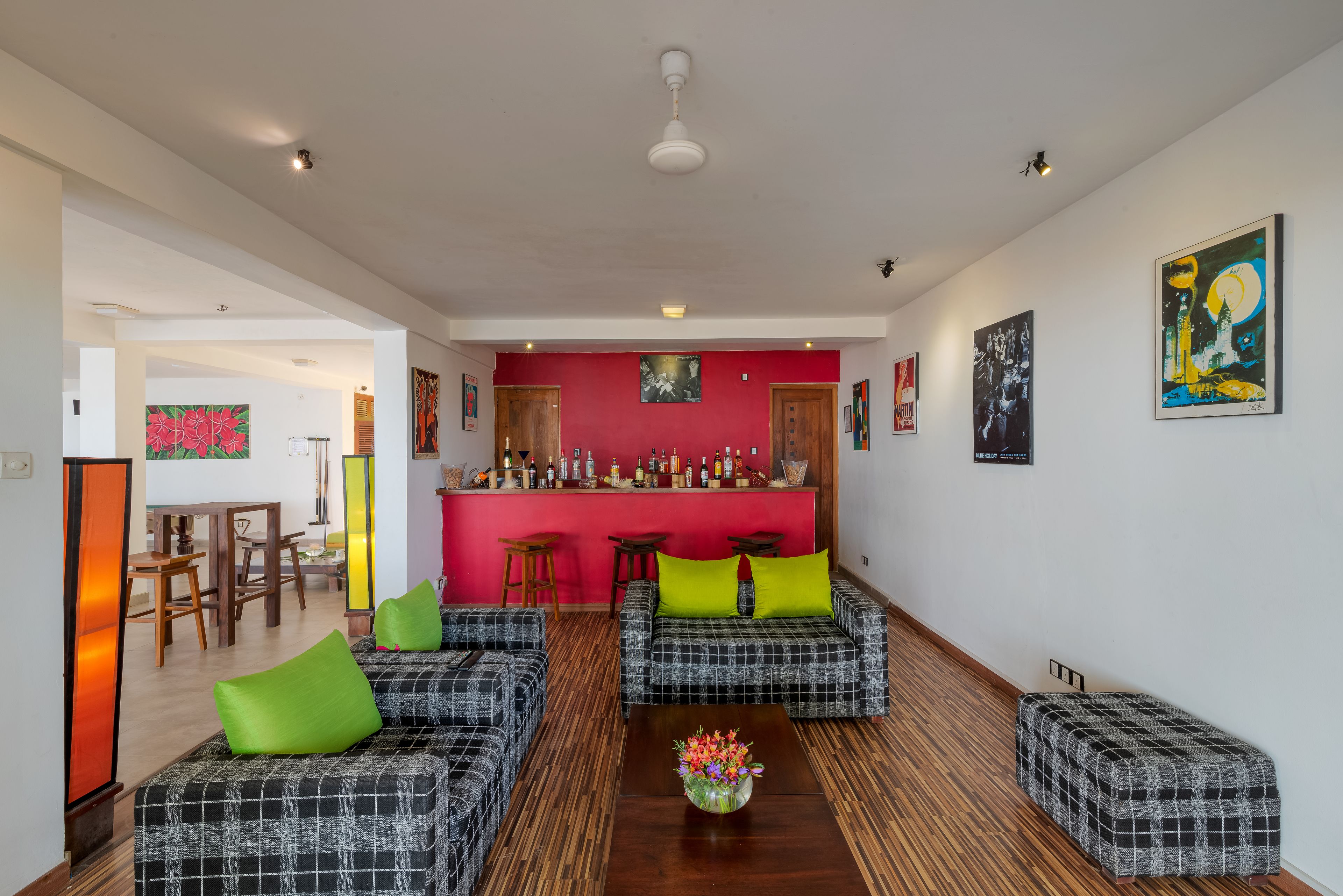 A modern living room with a red accent wall, two plaid sofas with green cushions, a wooden coffee table with a floral centerpiece, and a bar area with stools.