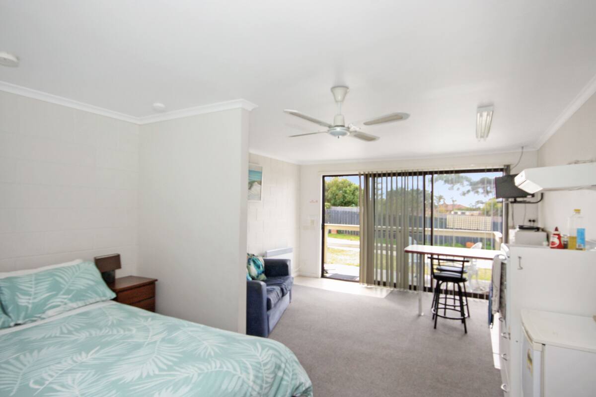 A bedroom with a bed, a desk, and a chair. There is a ceiling fan and a window with blinds.