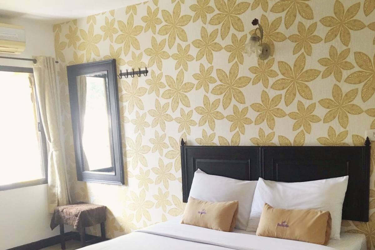 Een hotelkamer met een groot bed, een raam met gordijnen, een klein tafeltje met een stoel en een wand met een bloemenpatroon.