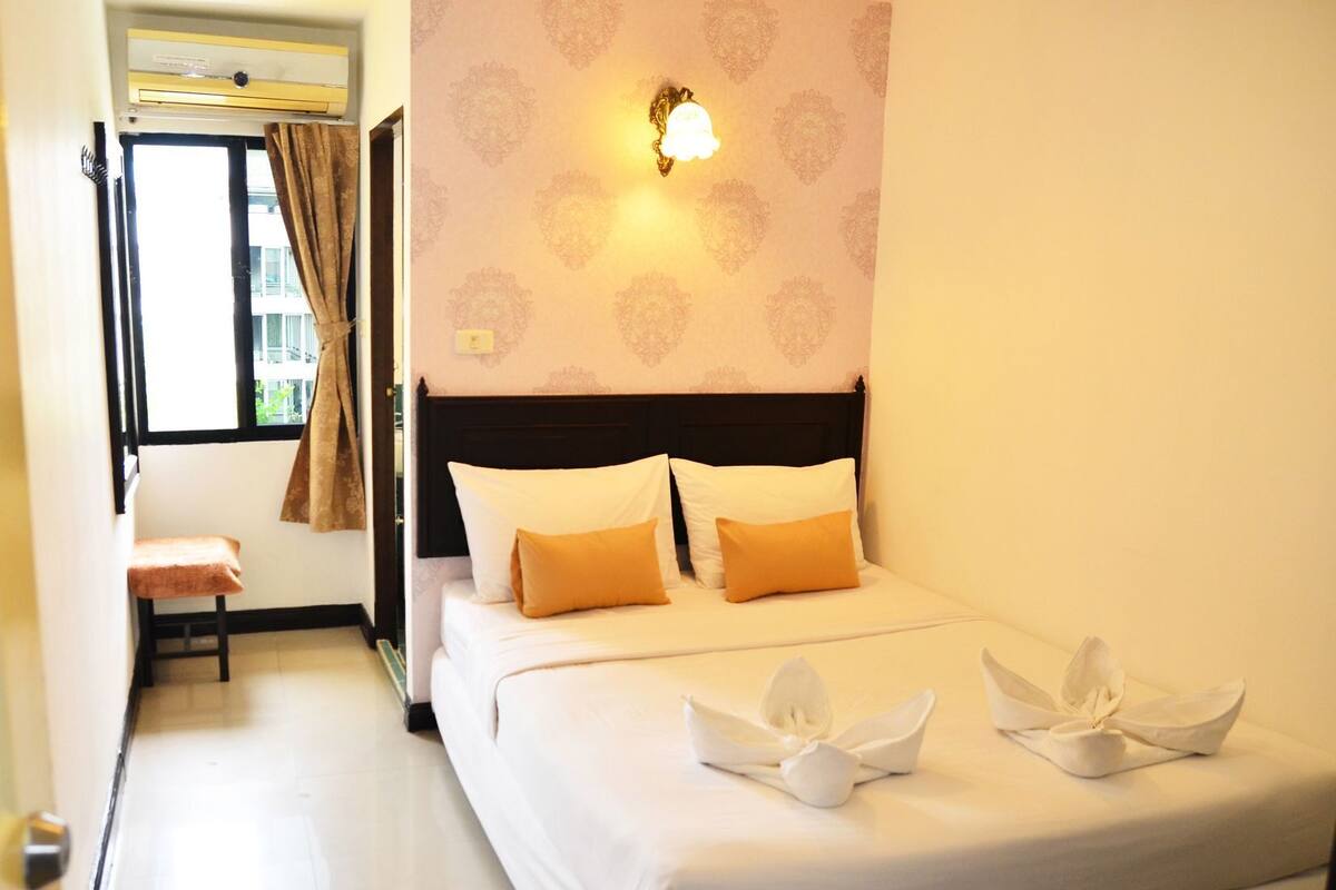 Een hotelkamer met een bed, een bankje, een raam met gordijnen en een wandlamp.