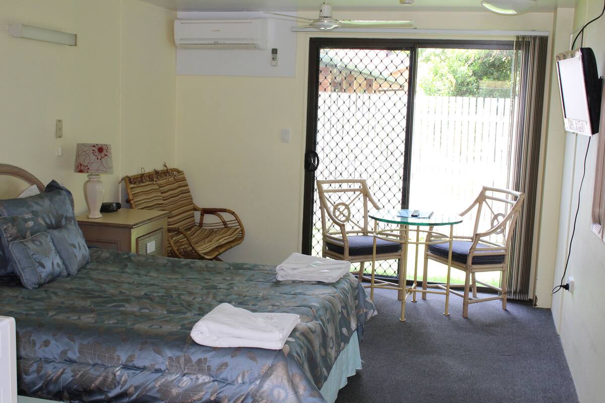 A hotel room with a bed, a desk with a lamp, a chair, a table, and a sliding glass door leading to an outdoor area.