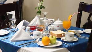 Una mesa de desayuno dispuesta con un florero, un vaso de jugo de naranja, tostadas, fruta y diferentes alimentos para el desayuno.