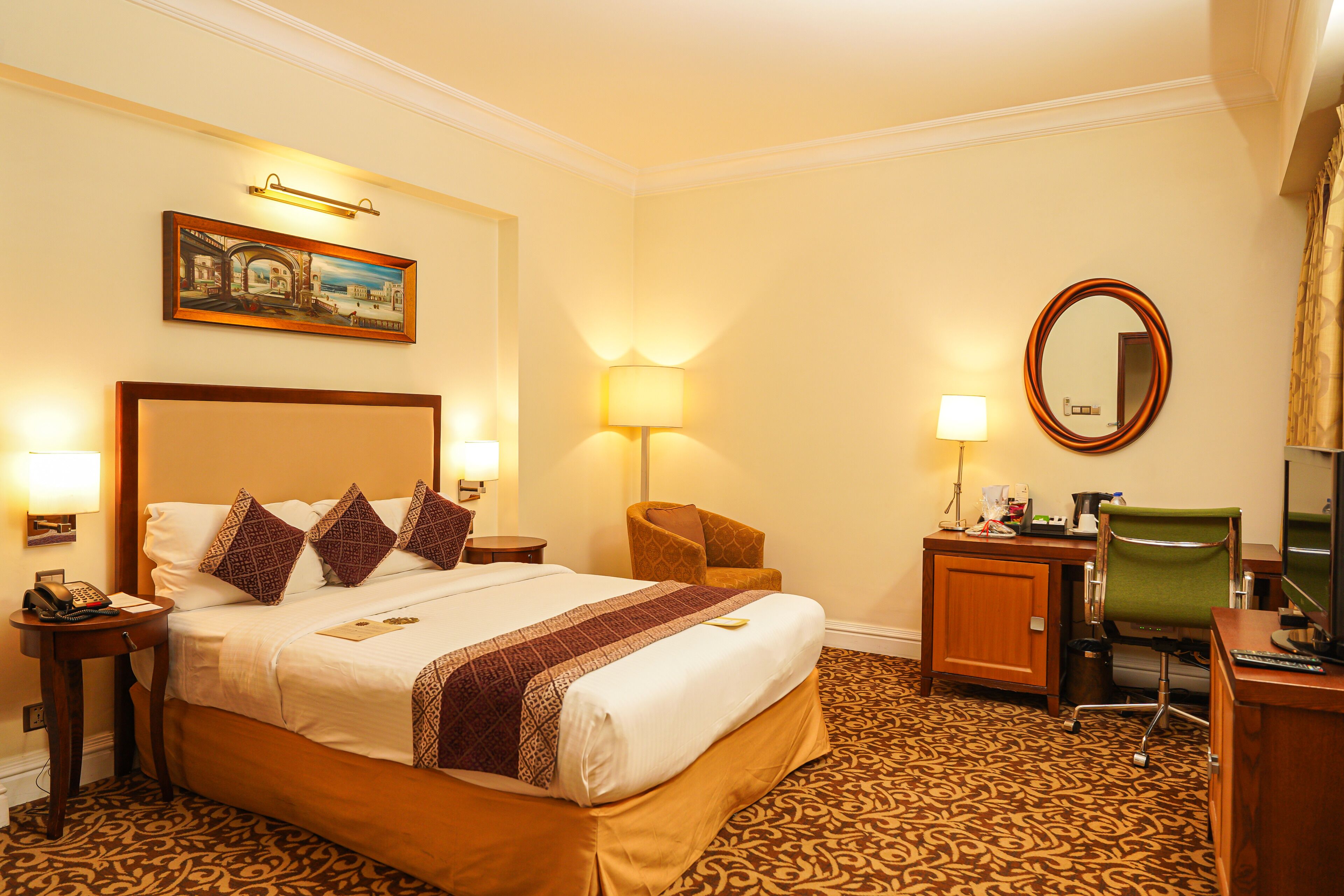 A hotel room with a large bed, a desk, a chair, a desk lamp, a telephone, and a painting on the wall.
