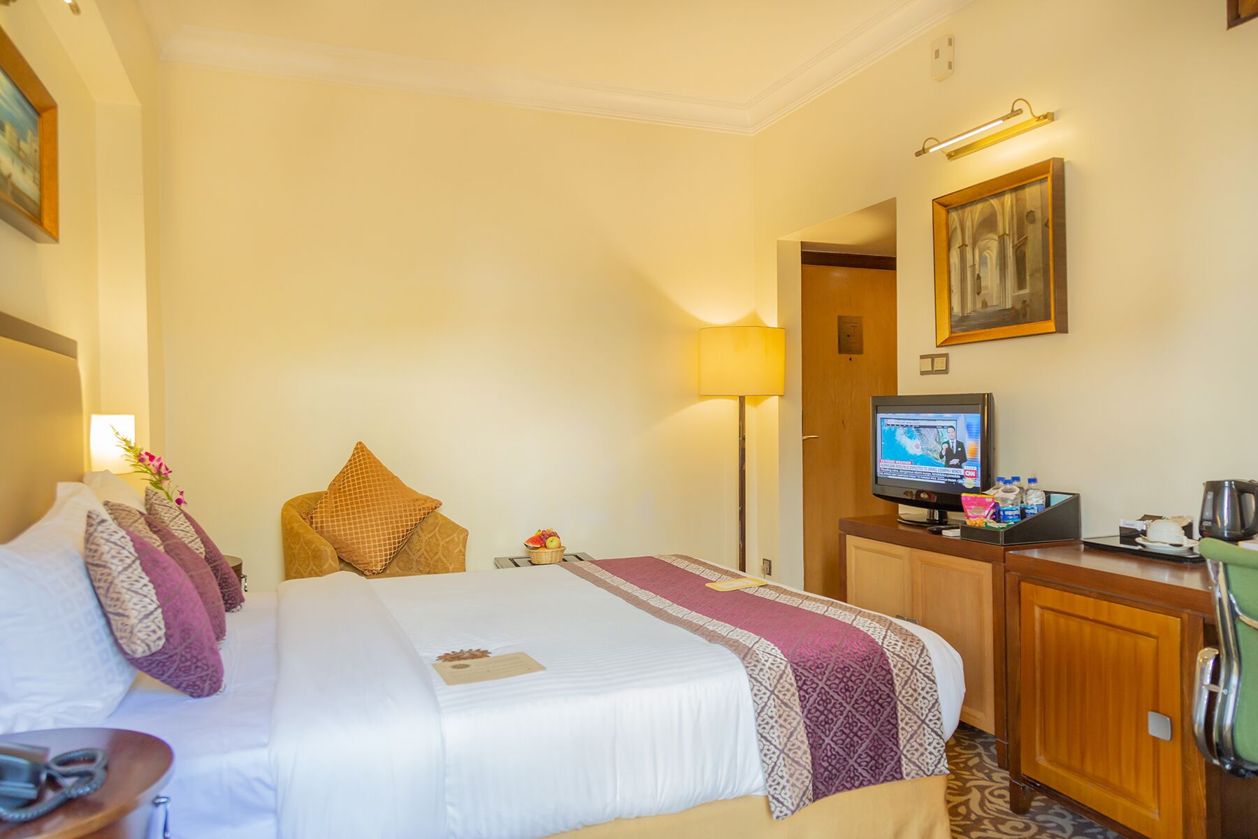 A hotel room with a bed, a desk with a television, a lamp, and a painting on the wall.