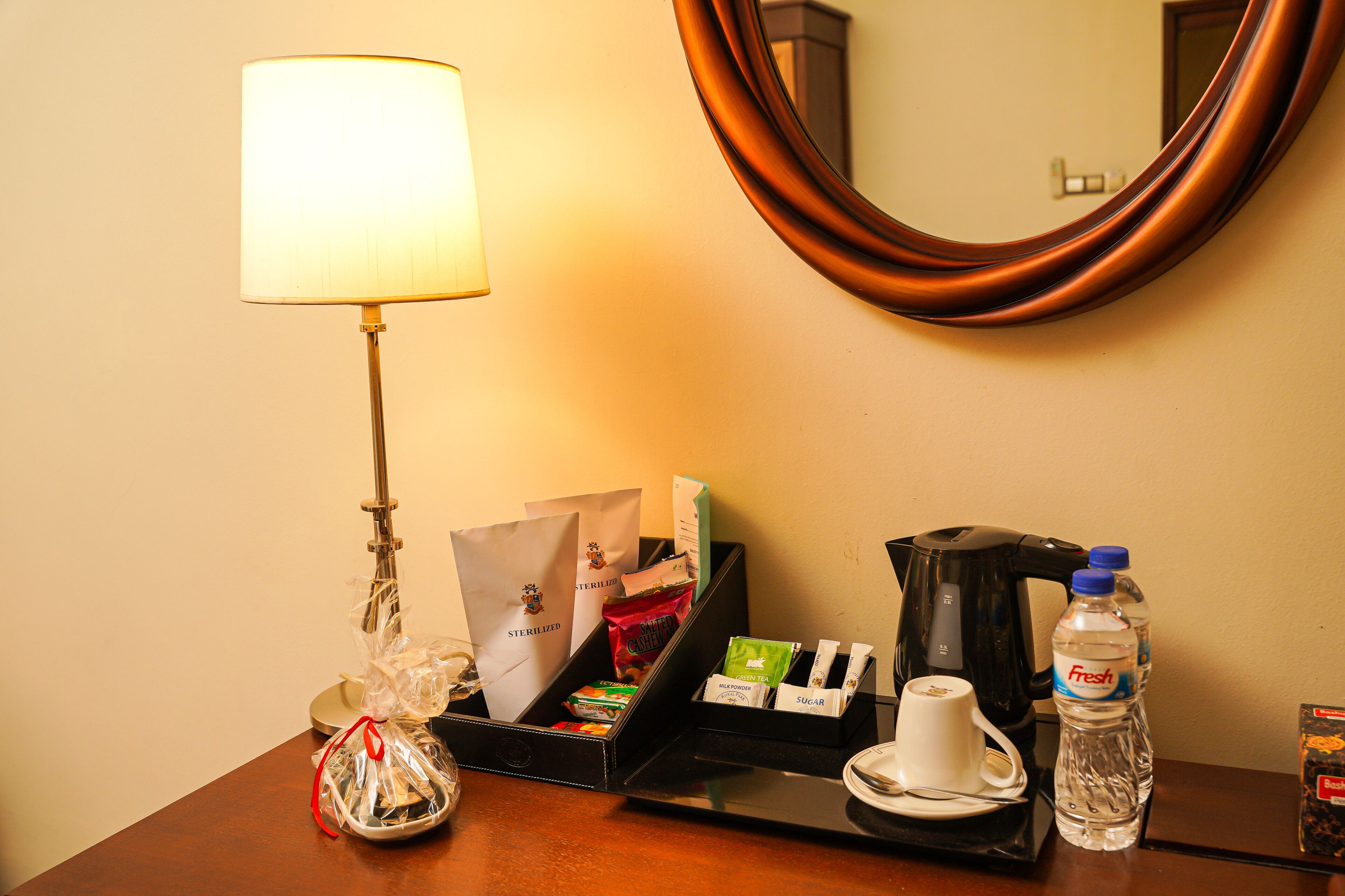 A hotel room with a bedside table featuring a lamp, a coffee maker, a water bottle, a cup, and a tray with various items including a bag and a box.